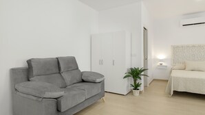 A modern bedroom with a grey sofa, a bed with a white headboard, a small bedside table with a lamp, and a potted plant.