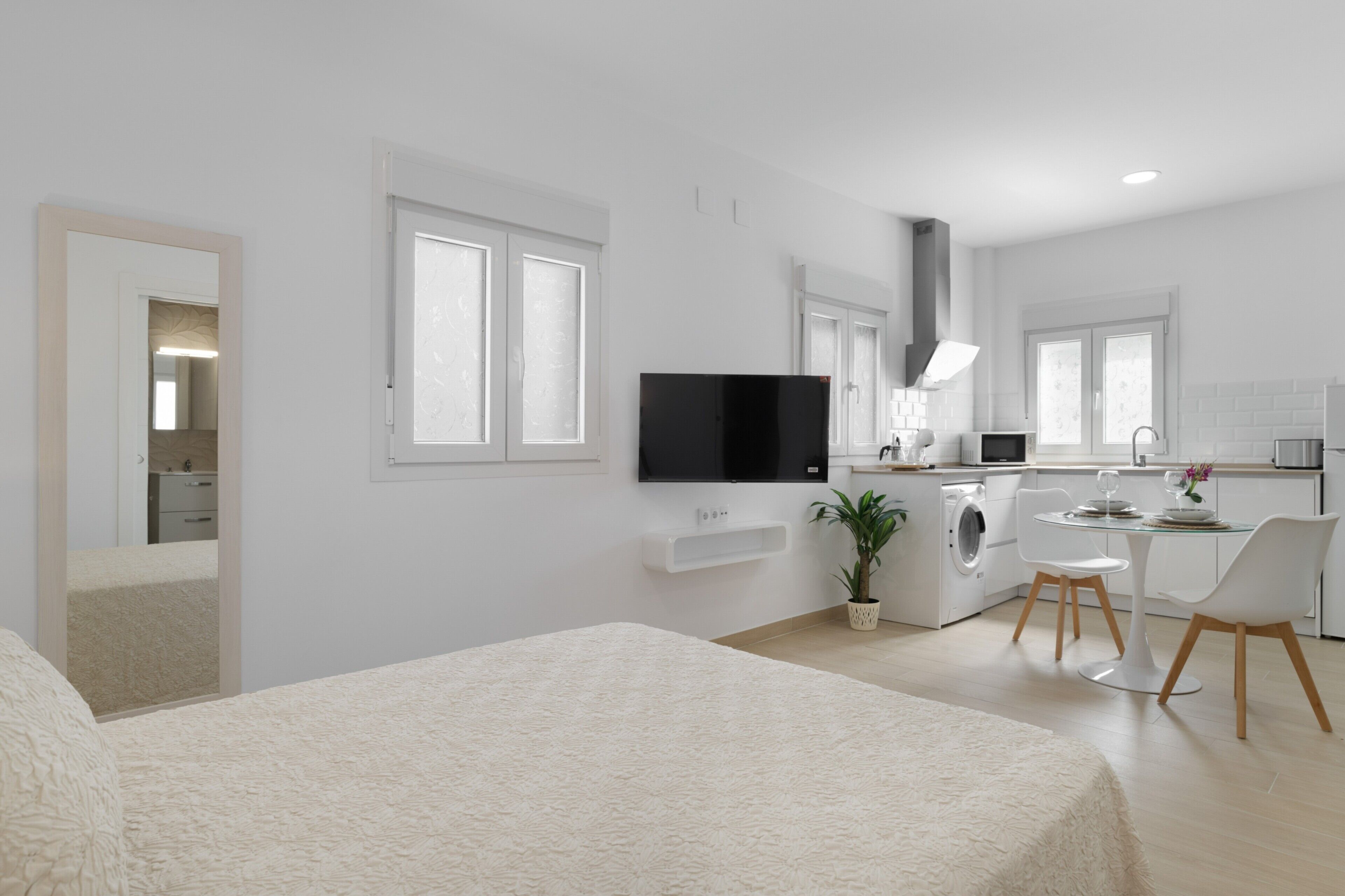 Ein moderner, gut beleuchteter Raum mit einem Bett, einem Fernseher, einer Essecke mit Stühlen und einer Küche inklusive Waschmaschine.