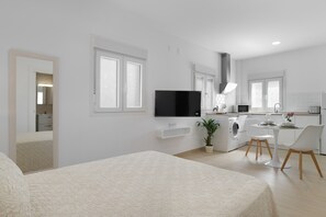 Una habitación moderna y bien iluminada con una cama, un televisor, un comedor con sillas y una cocina con lavadora.