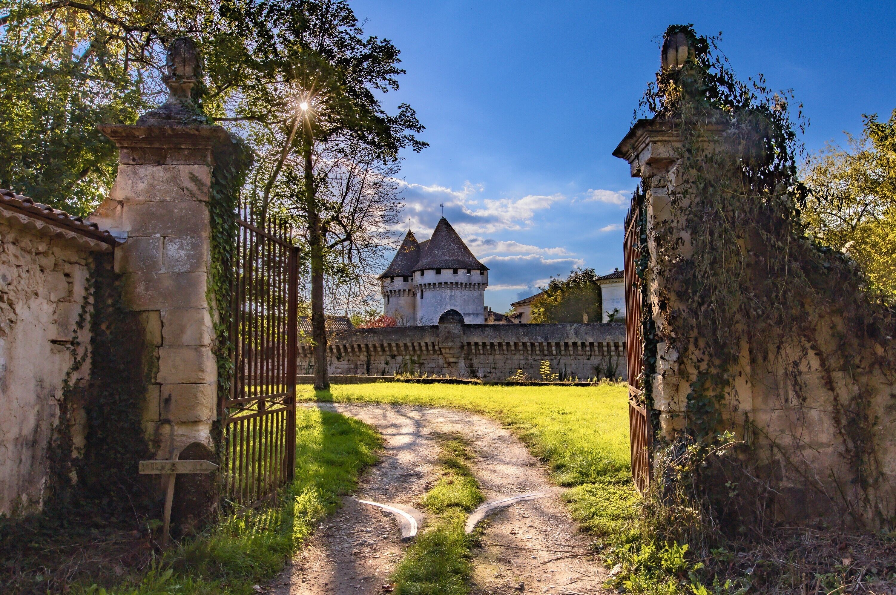 Ein Schlossportal mit einem Weg, der zu einem Turm führt, umgeben von Grünflächen und Steinmauern.