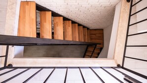 Un escalier étroit avec des marches en bois et une rampe métallique, menant à une mezzanine dotée d’un plafond en pente et de murs en briques apparentes.