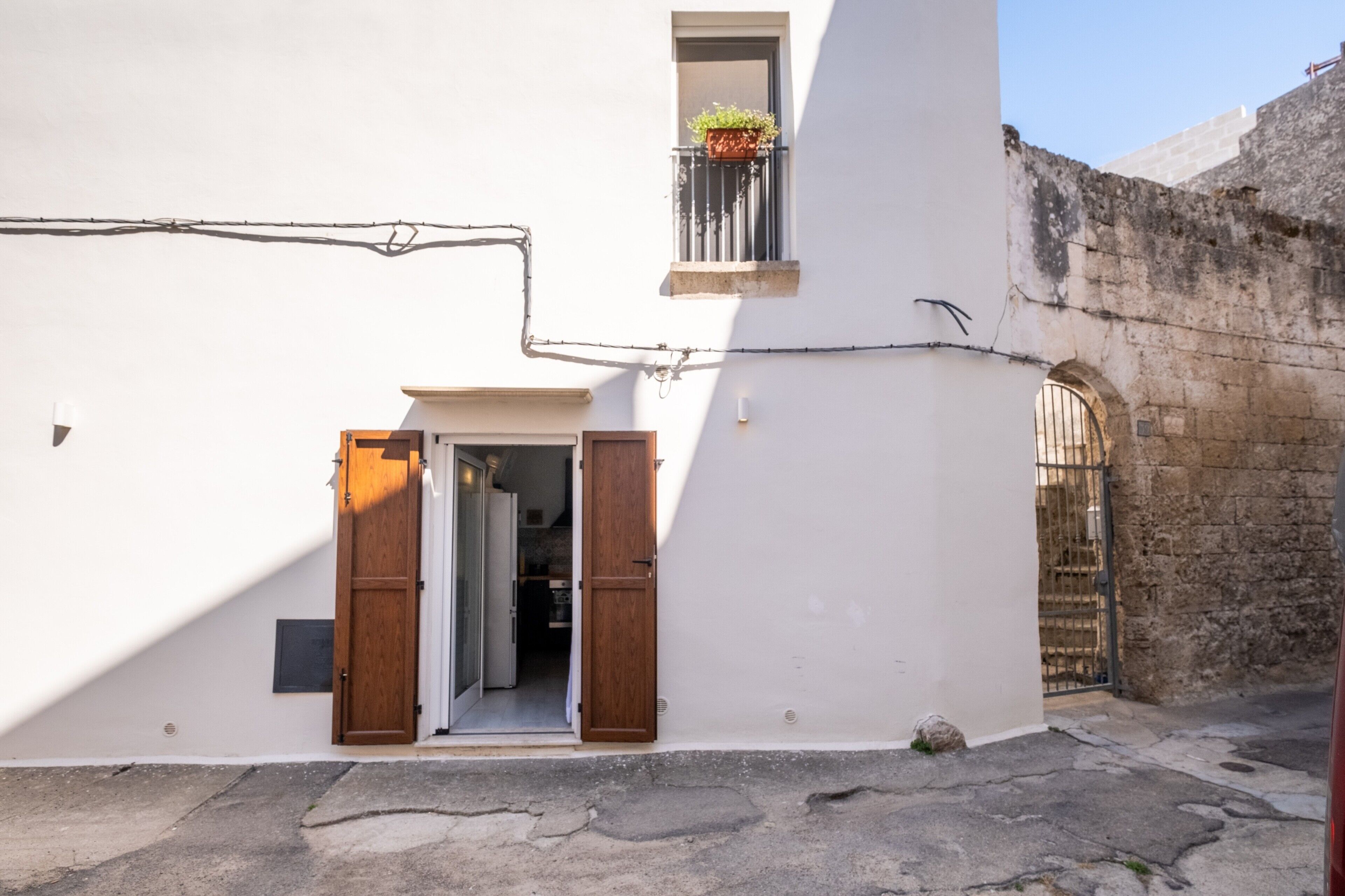 Una calle estrecha con un edificio que presenta una puerta de madera, un pequeño ventanal con una planta y un muro de piedra.