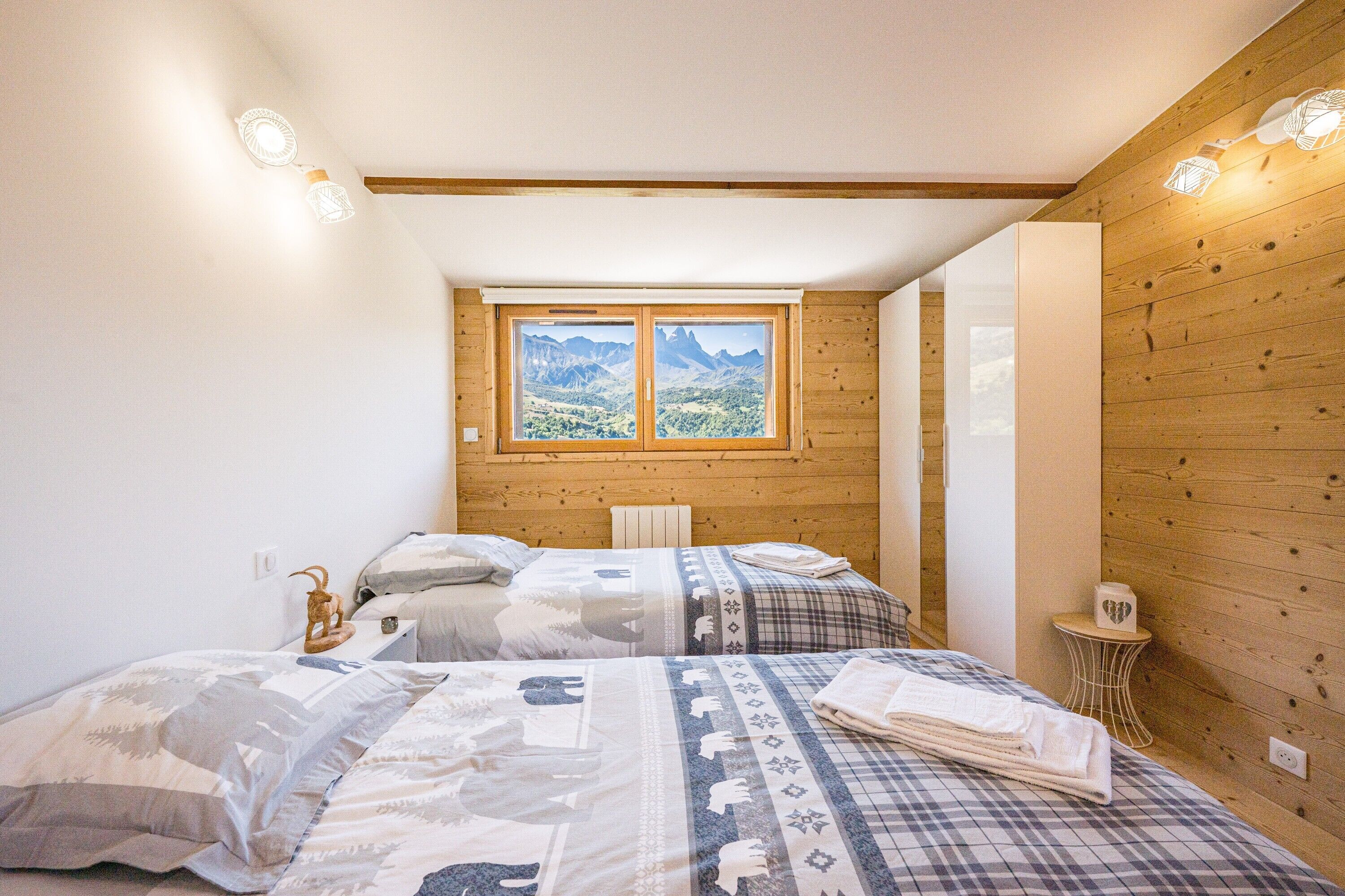 Un dormitorio con dos camas, una ventana con vistas a la montaña, paredes de madera y una mesita de noche con una lámpara.