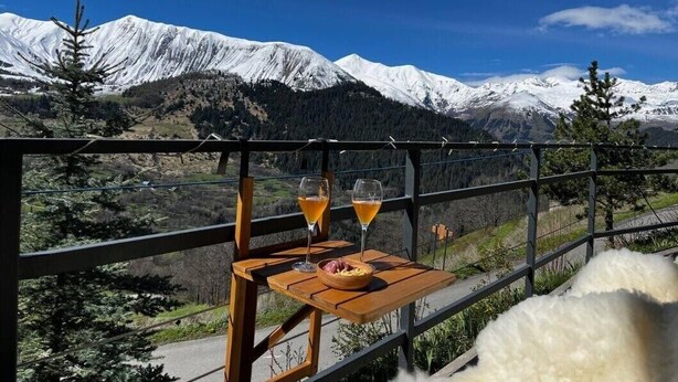 Ein Balkon mit Tisch, zwei Gläsern eines gelben Getränks und einer Schüssel Essen, mit Blick auf eine verschneite Berglandschaft.