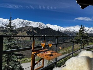 Un balcon avec une table, deux verres contenant une boisson jaune et un bol de nourriture, donnant sur un paysage montagneux enneigé.