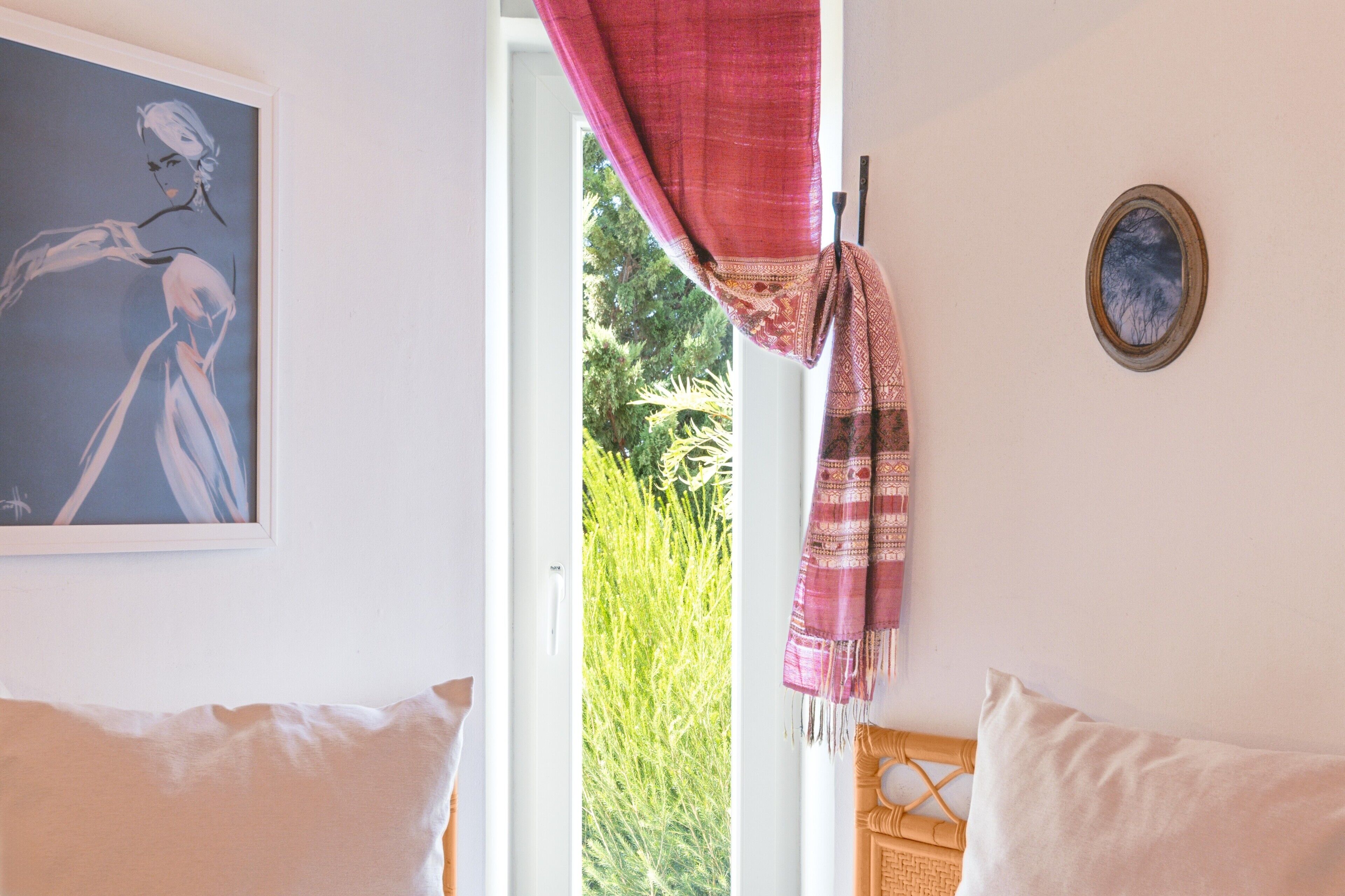A room with a bed, a window with a view of greenery, a painting on the wall, and a decorative item on the wall.
