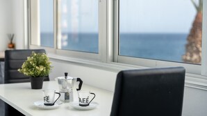 A table with a coffee maker, two cups, and a potted plant by a window overlooking the sea.