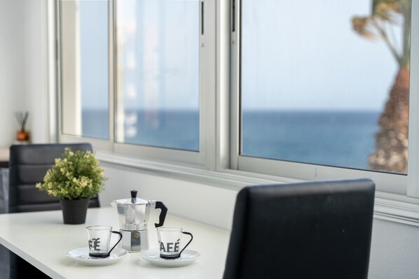 Uma mesa com uma cafeteira, duas xícaras e um vaso de planta, próxima a uma janela com vista para o mar.
