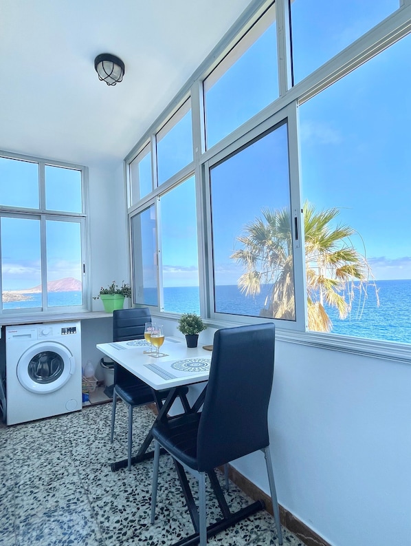 A room with a washing machine, a table with chairs, and large windows overlooking the sea.