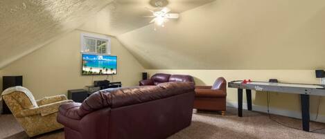 A cozy attic room with a brown leather sofa, a small armchair, a pool table, and a ceiling fan.