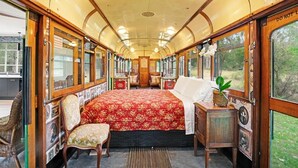 A vintage train interior with a central bed, wooden furniture, and large windows offering views of the outdoors. - Seclusion, Tram, Native Bush, Wineries, Beaches, Melbourne Icon, Village nearby (Balnarring)