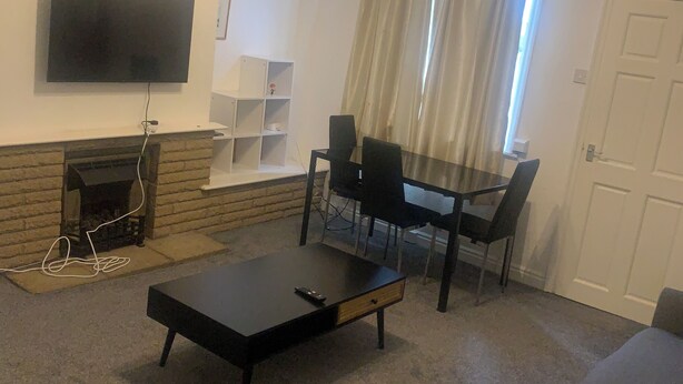 Een woonkamer met een flatscreen televisie aan de muur, een open haard, een salontafel en een eethoek met stoelen en een tafel.