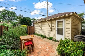 Una piccola casa con patio in mattoni, un tavolo e una sedia di legno e un giardino con diverse piante.