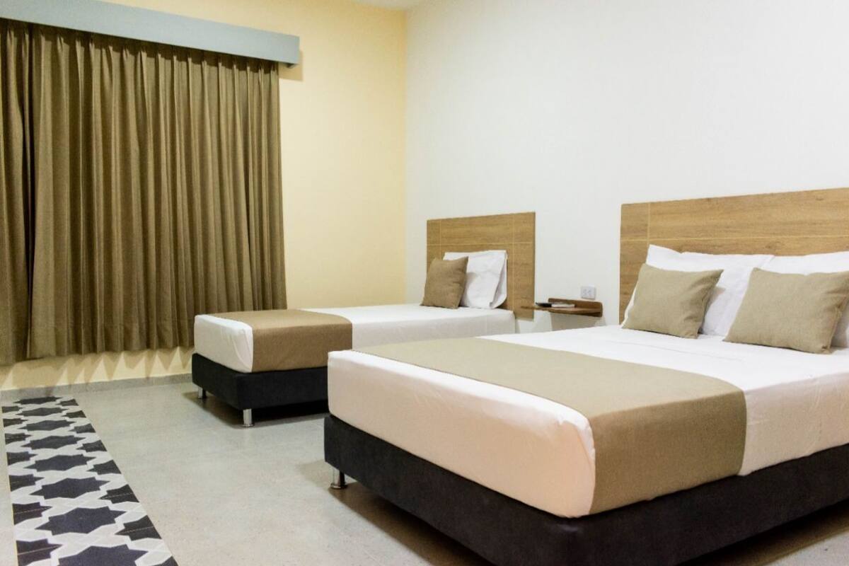 Quarto de hotel com duas camas, cabeceira de madeira, mesa de cabeceira e tapete estampado.