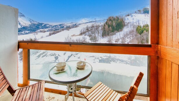 Un balcon avec une table et des chaises, donnant sur un paysage enneigé avec des montagnes et une rivière.
