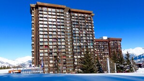 Un hôtel à plusieurs étages doté de balcons, situé près d’une étendue d’eau et de montagnes enneigées.