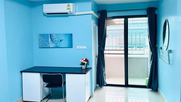 A room with a desk, chair, air conditioning unit, a painting, a flower vase, and a sliding glass door to a balcony.