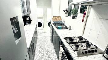 A kitchen with a stainless steel refrigerator, a stove, and a sink. There is a patterned floor and a door leading to another room.