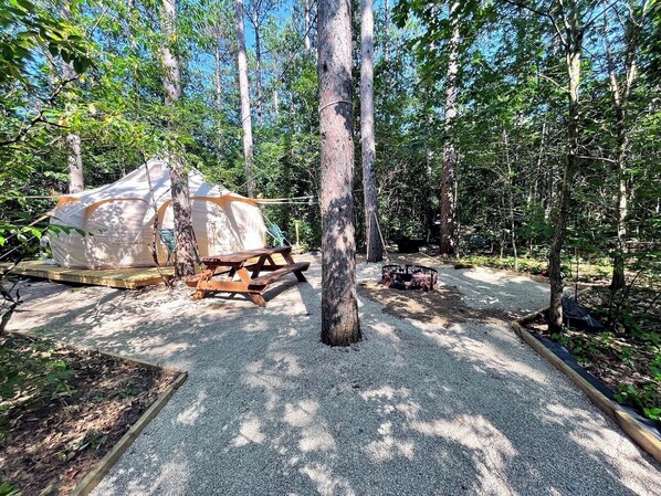 Un terrain de camping situé dans une forêt, équipé d’une grande tente, d’une table de pique-nique et d’un foyer.