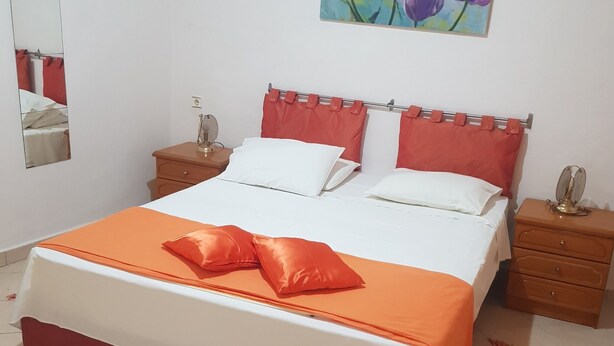 A neatly made bed with a red and white bedspread, two white pillows, and an orange decorative pillow. There is a wooden nightstand with a small lamp and a mirror on the wall beside the bed. - Maria House in the port of Kalymnos (Kalymnos)