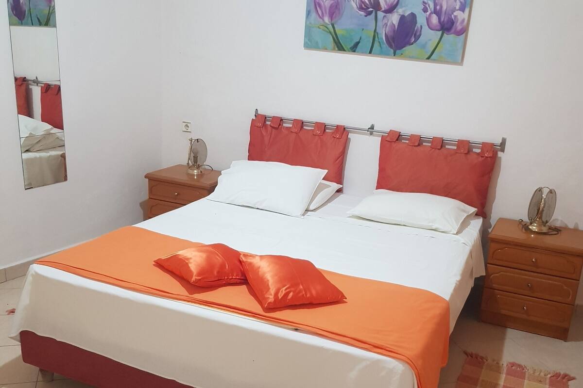 A neatly made bed with a red and white bedspread, two white pillows, and an orange decorative pillow. There is a wooden nightstand with a small lamp and a mirror on the wall beside the bed.