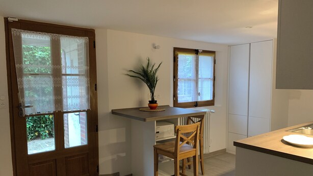 Une cuisine avec une porte en bois, une fenêtre ornée de rideaux en dentelle, une plante en pot et une chaise.
