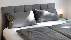 A bed with grey bedding, a grey upholstered headboard, and a bedside table with a remote control and a lit lamp.
