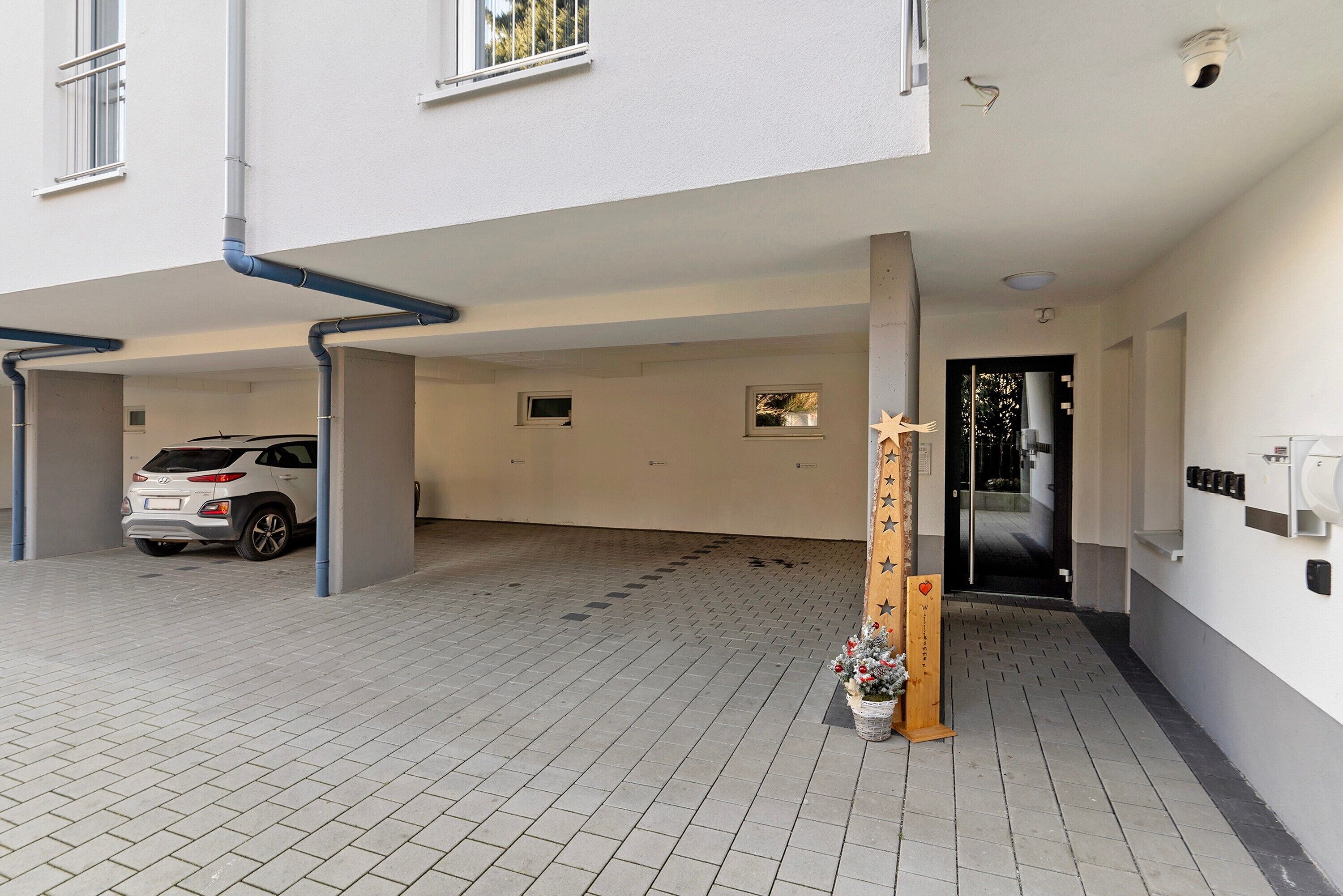 Une entrée d’immeuble avec un espace de stationnement couvert, une voiture blanche et une structure décorative en bois agrémentée de fleurs.