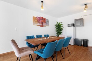 A dining area with a wooden table, blue chairs, a painting, a potted plant, and a microwave.