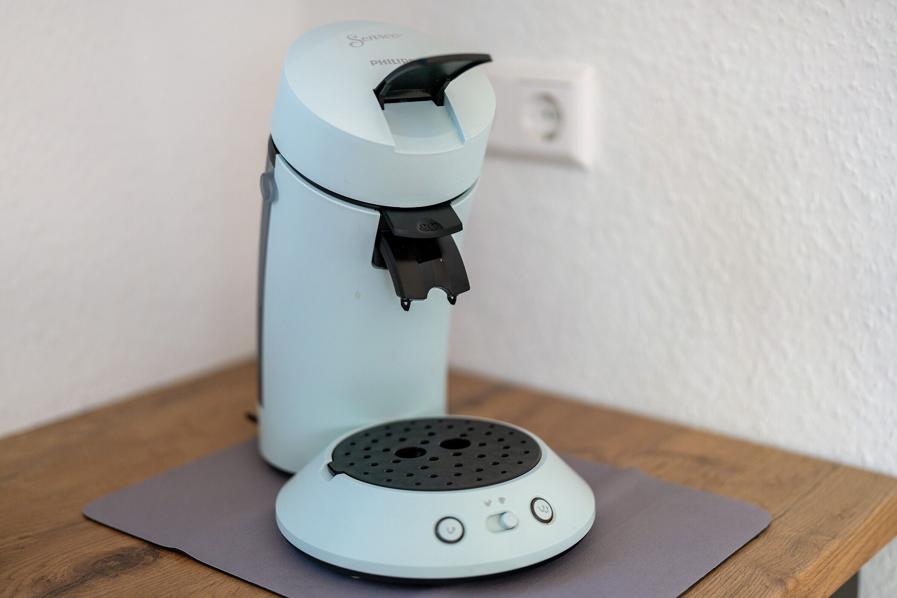 Eine Kaffeemaschine auf einem Holztisch mit einer grauen Unterlage, eine Steckdose im Hintergrund und eine weiße Wand.