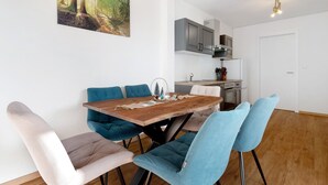 A dining area with a wooden table and chairs, a kitchen in the background, and a painting on the wall.