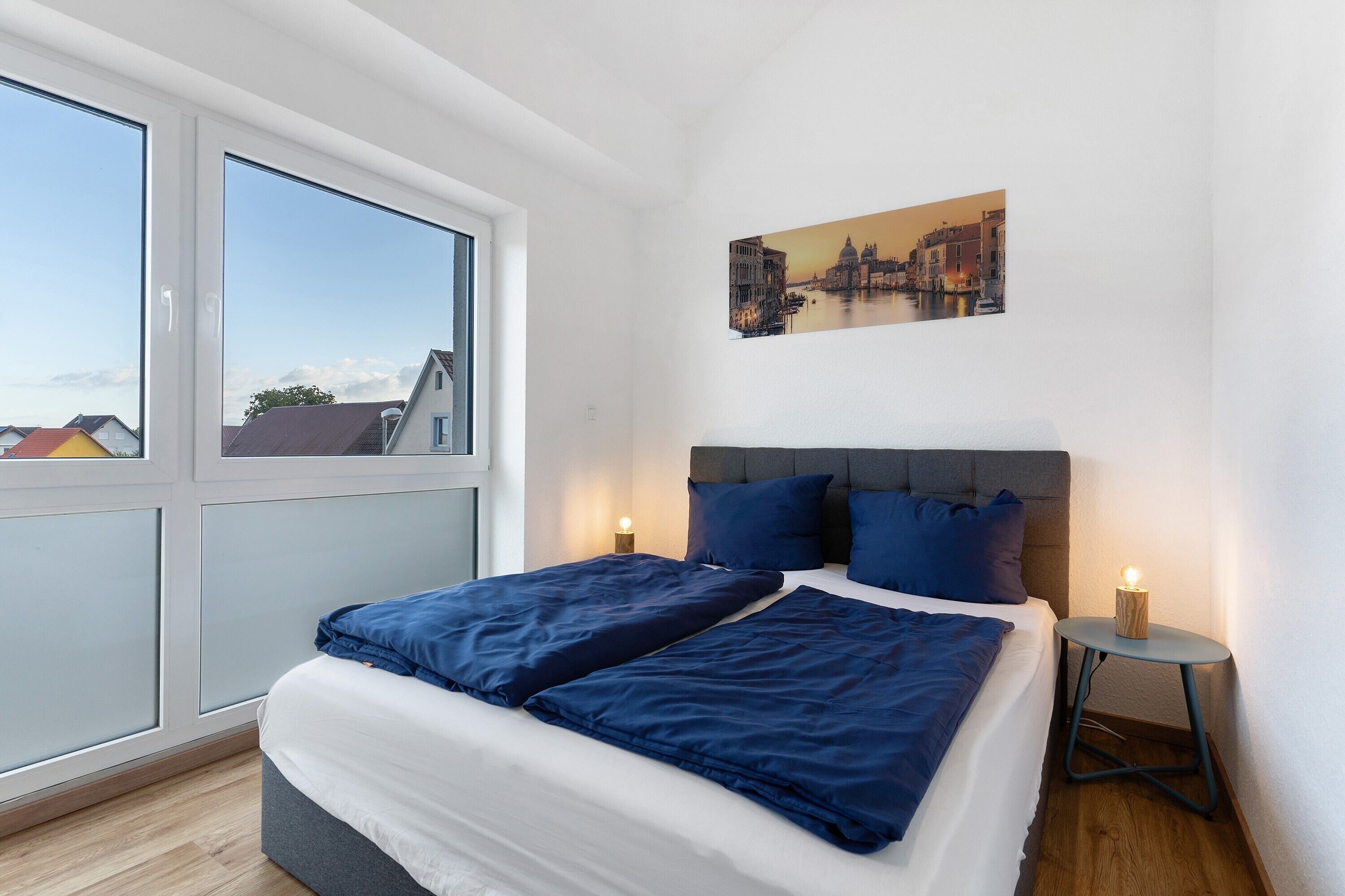A bedroom with a bed, a nightstand, a wall-mounted painting, and a window.