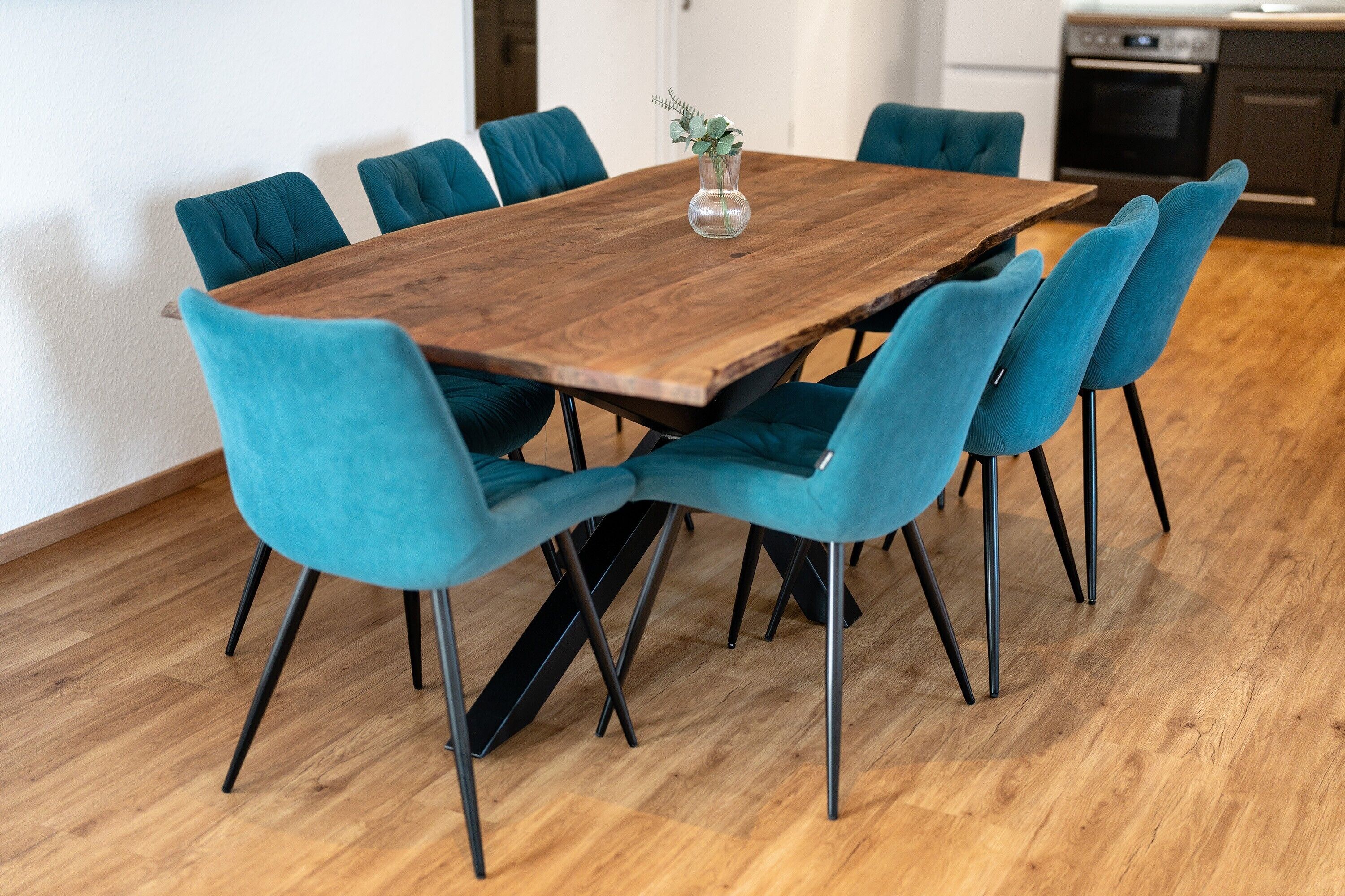 A wooden dining table with six teal chairs, a vase with a plant, and a kitchen in the background.