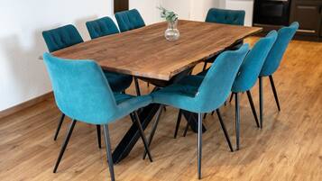 A wooden dining table with six teal chairs, a vase with a plant, and a kitchen in the background.