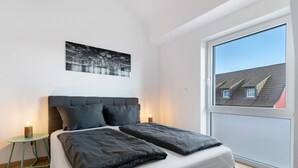 A modern bedroom with a large bed, a cityscape painting, a small side table, and a large window.