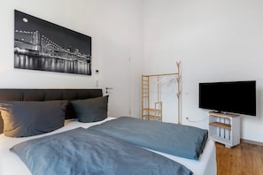 A bedroom with a bed, pillows, a TV, a coat rack, and a painting of a cityscape.