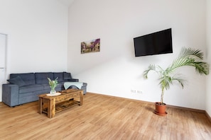 Ein Wohnzimmer mit grauem Sofa, einem Holztisch, einem Blumentopf, einem Flachbildfernseher und einem an der Wand hängenden Bild.
