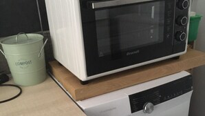 Une petite cuisine équipée d’un micro-ondes posé sur le dessus d’un lave-linge, d’un bac à compost et d’une planche à découper en bois.