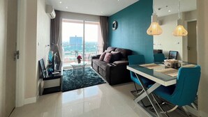 A modern living room with a sofa, a dining table with chairs, a view of the city through large windows, and a balcony with a glass table and chairs. - Grande Carribean Condo By Thita (Pattaya)