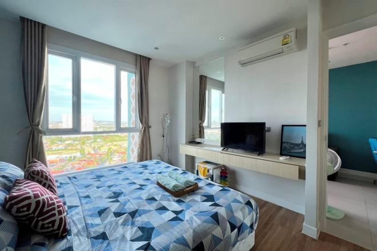 Une chambre d’hôtel avec un grand lit, une télévision, un bureau avec un ordinateur et une vue sur la ville grâce à de grandes fenêtres.
