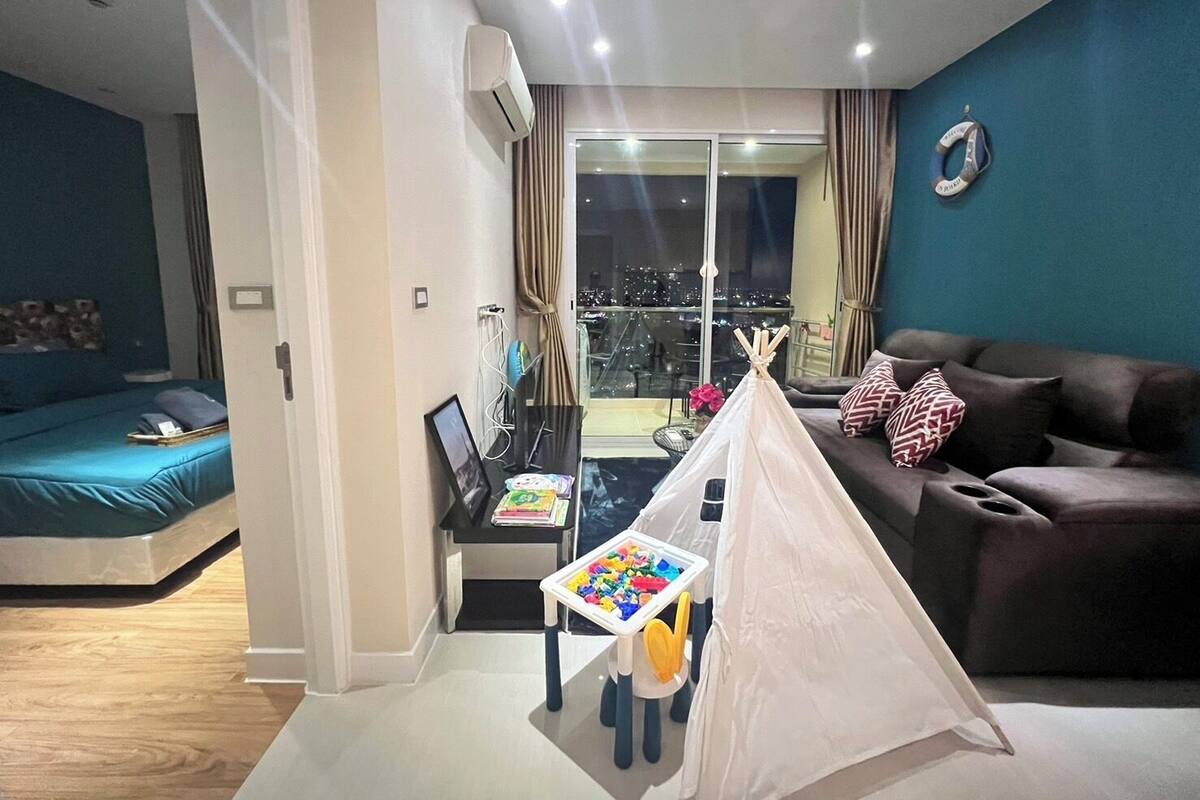 A modern living room with a sofa, a small table with toys, a tent, and a city view through large windows.