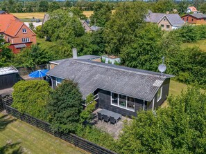A modern house with a dark roof, surrounded by lush greenery and other houses in the background.