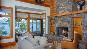 A spacious living room with a stone fireplace, large windows offering a view of a lake, and a cozy seating area.