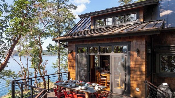 A modern cabin with a deck overlooking a lake, featuring a dining area with red chairs and a table set for four.
