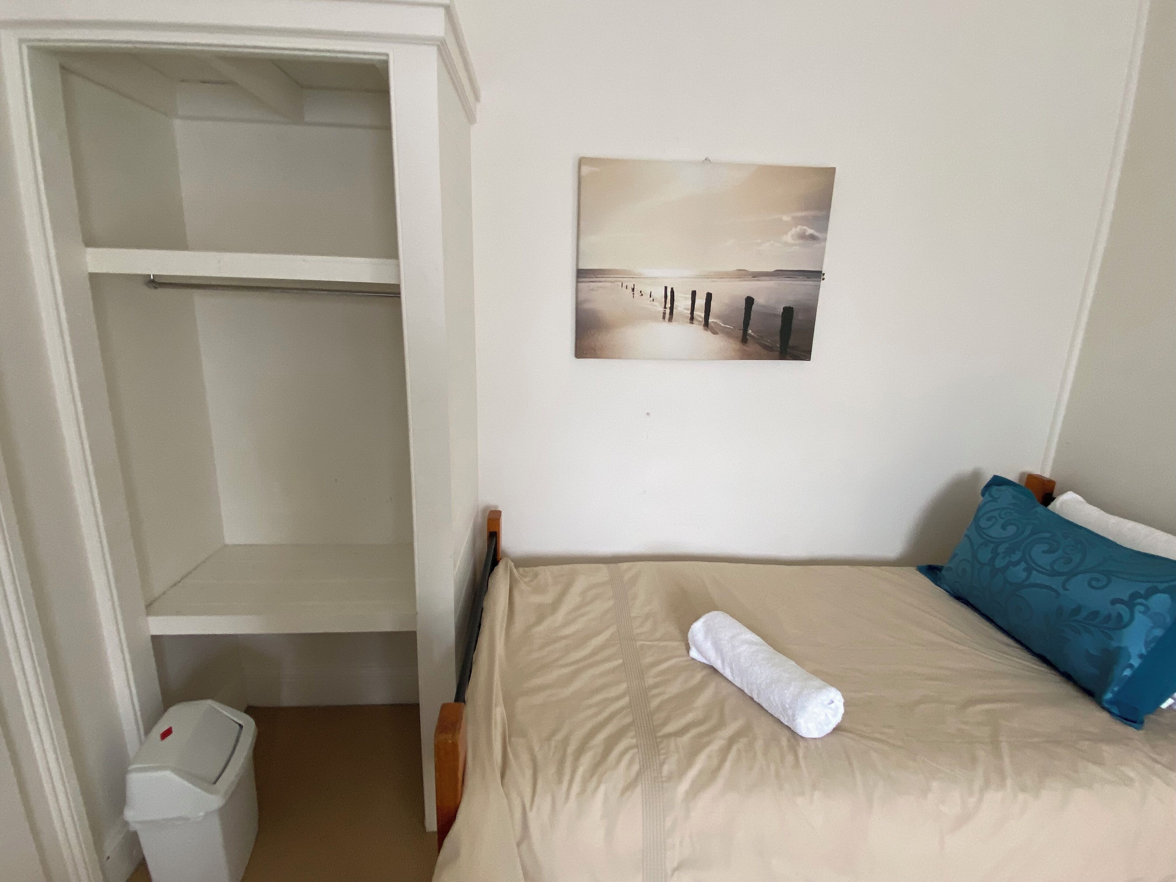 A bedroom with a bed, a white wardrobe, a picture on the wall, and a trash bin.