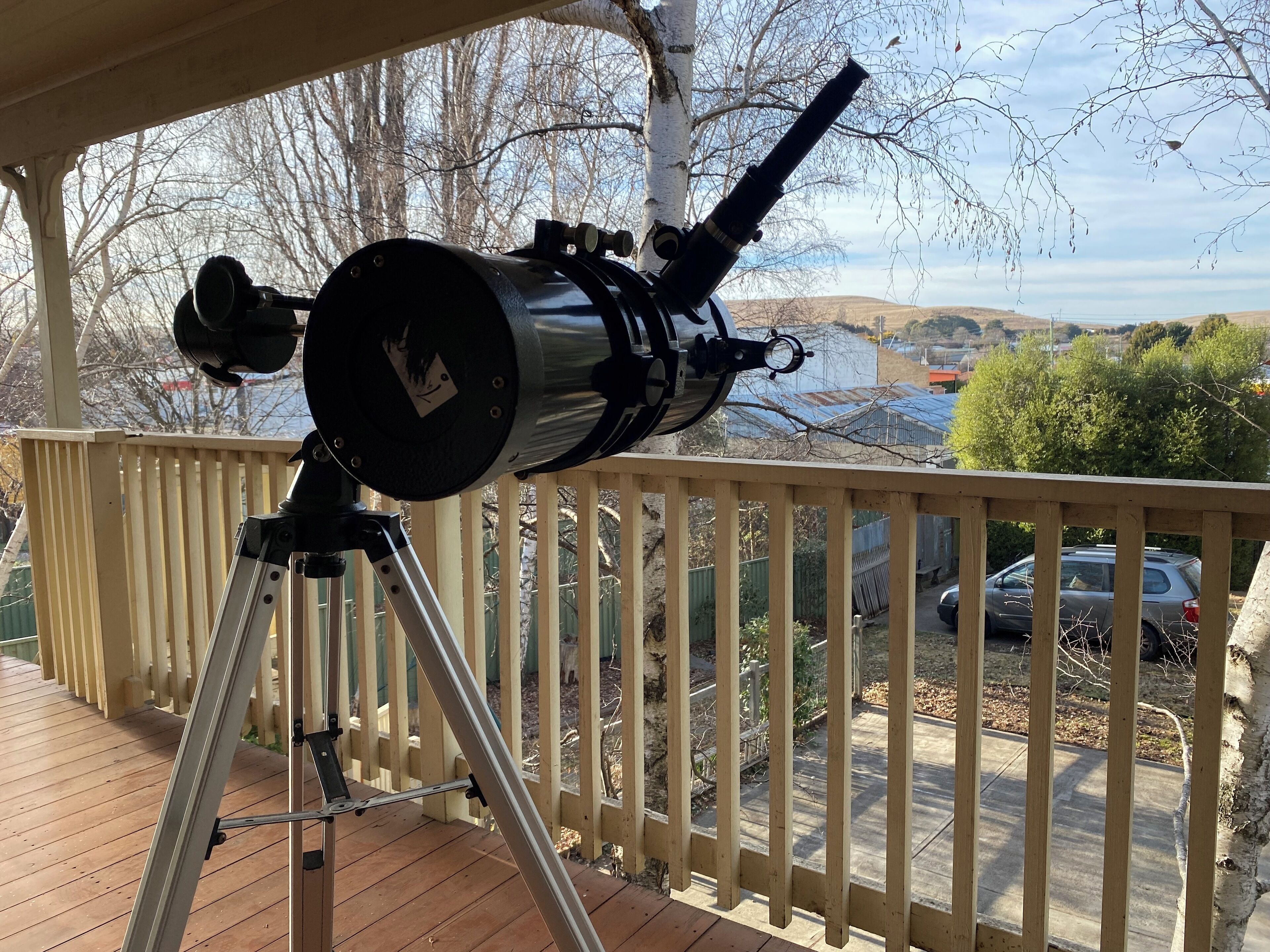 A telescope on a wooden deck with a railing, overlooking a residential area with houses and trees.