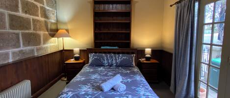Un dormitorio con una cama grande, dos mesitas de noche con lámparas, cabecera de madera, pared de piedra, radiador y ventana con cortinas.