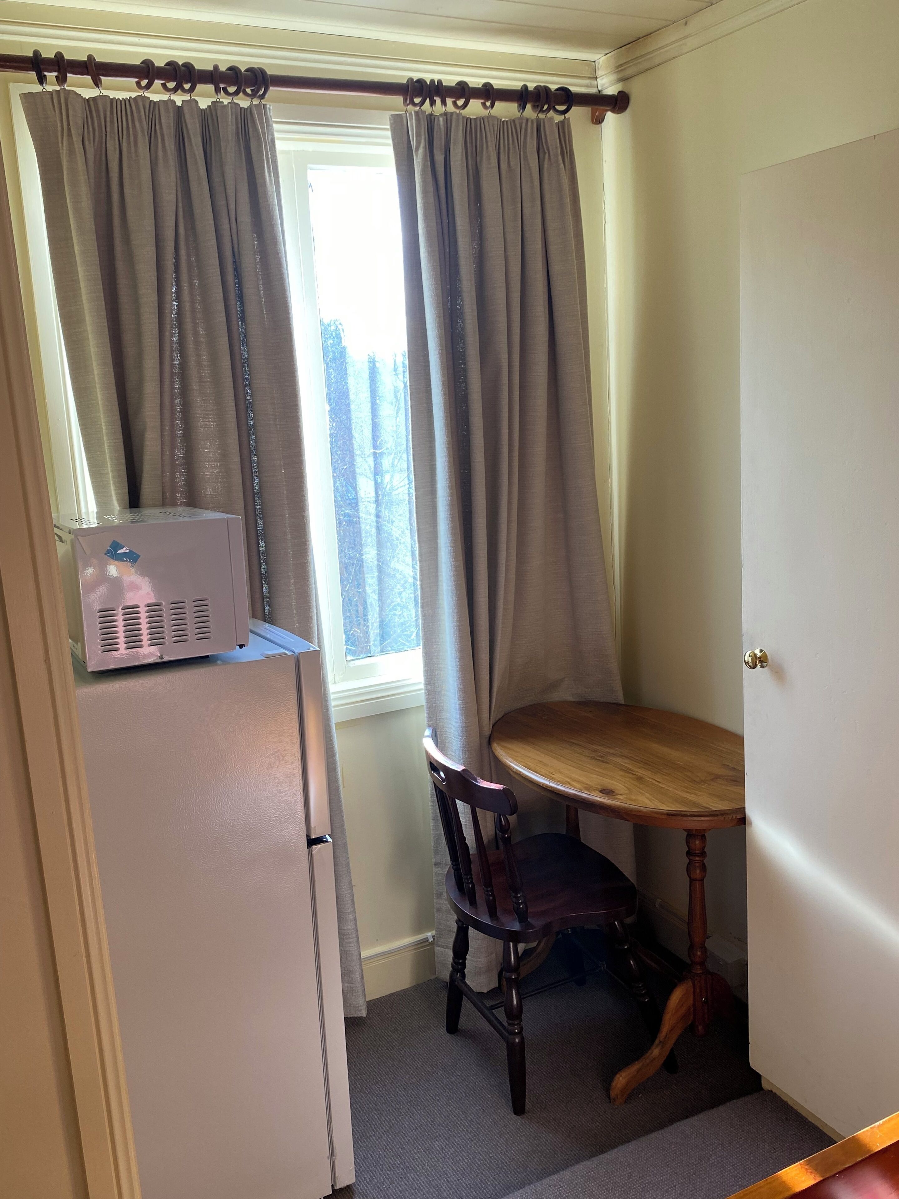 A corner room with a window, a table, a chair, and a refrigerator.