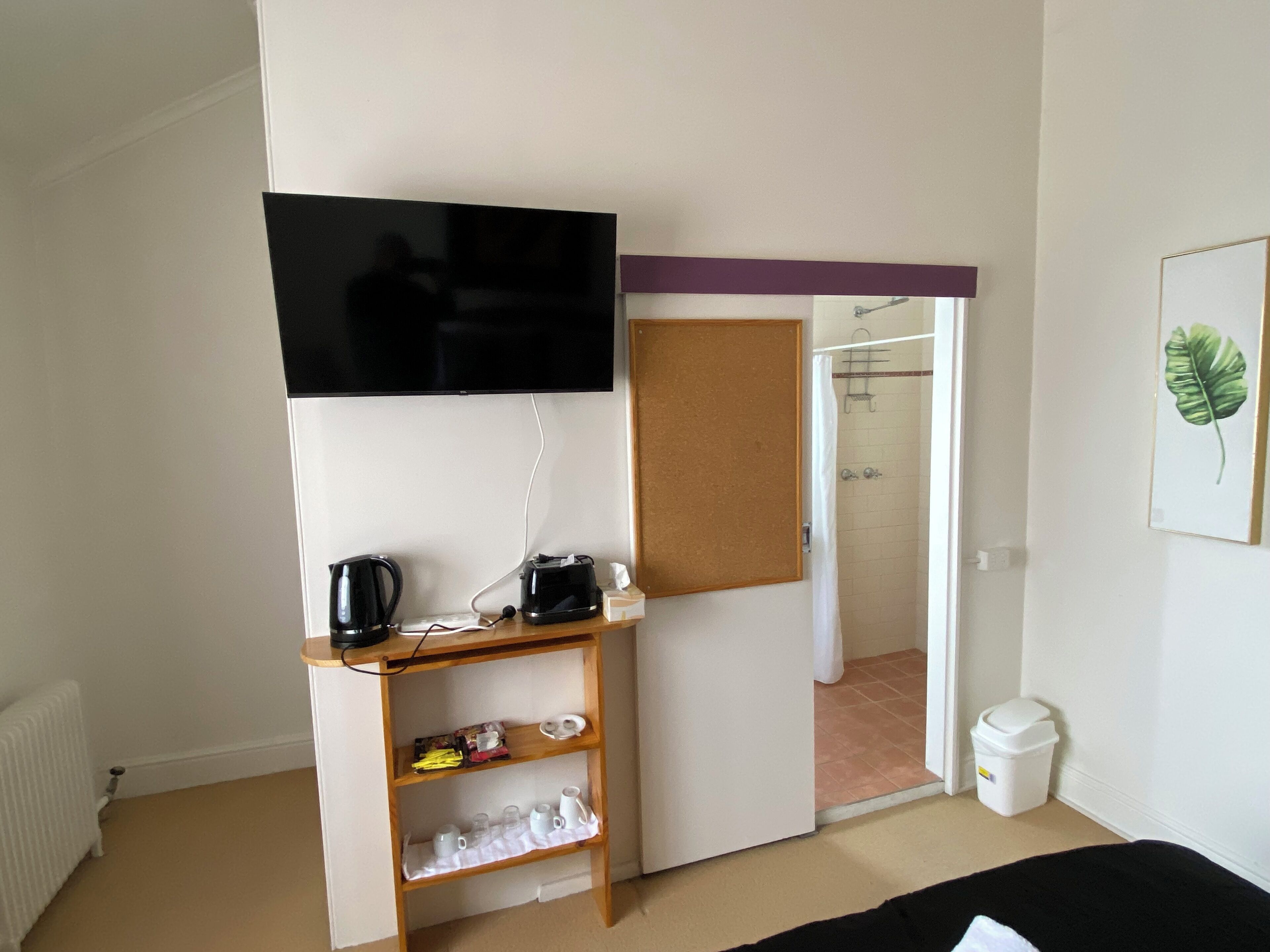 A bedroom with a bed, a TV mounted on the wall, a shelf with various items, a door leading to a bathroom, and a trash bin.