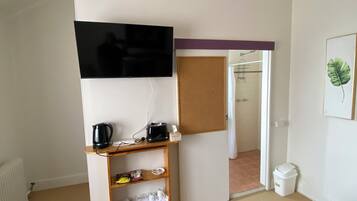 A bedroom with a bed, a TV mounted on the wall, a shelf with various items, a door leading to a bathroom, and a trash bin.